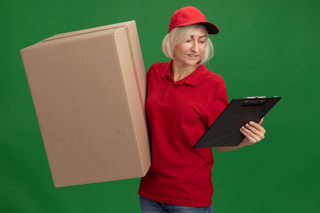 Lächelnde blonde Lieferfrau mittleren Alters in roter Uniform und Mütze mit Karton und Zwischenablage mit Blick auf die Zwischenablage isoliert auf grüner Wand