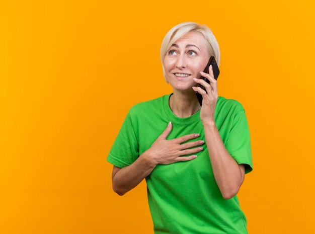 Lächelnde blonde slawische Frau mittleren Alters, die am Telefon spricht und Hand auf Brust hält, die Seite lokalisiert auf gelber Wand mit Kopienraum betrachtet