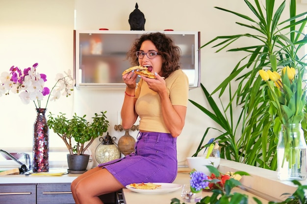 Lächelnde brünette Frau mit lockigem Haar sitzt auf einem Boden und isst Pizza in der Küche.