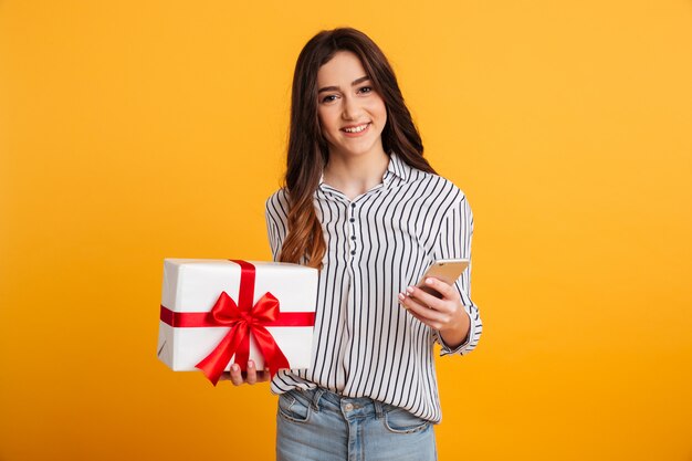 Lächelnde Brunettefrau im Hemd, das Geschenkbox und Smartphone hält