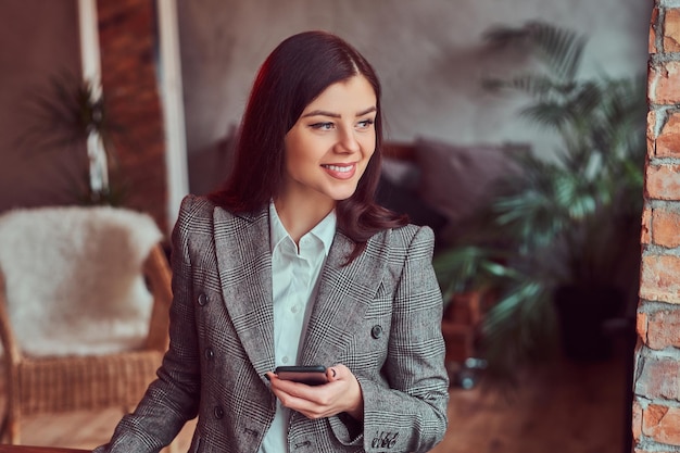 Lächelnde charmante brünette Frau in einer grauen, eleganten Jacke, die ein Smartphone in einem Raum mit Loft-Interieur hält und wegschaut.