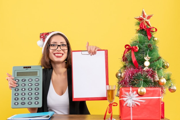 Lächelnde charmante Dame im Anzug mit Weihnachtsmannhut, der Dokument zeigt und Rechner im Büro auf Gelb lokalisiert hält