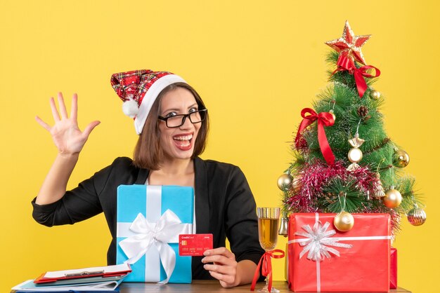 Lächelnde charmante Dame im Anzug mit Weihnachtsmannhut und Brille, die fünf zeigt und Geschenk und Bankkarte im Büro auf gelb lokalisiert hält