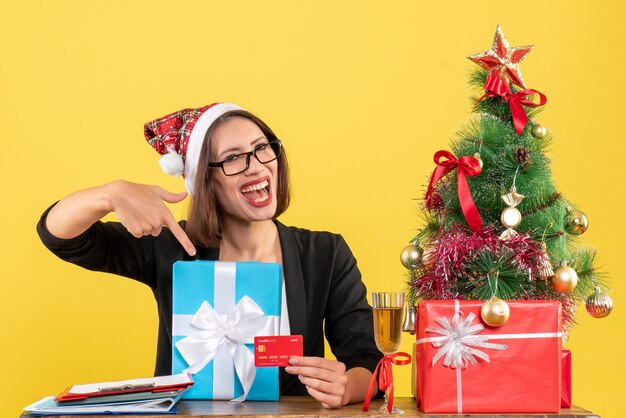 Lächelnde charmante Dame im Anzug mit Weihnachtsmannhut und Brille, die Geschenk und Bankkarte im Büro auf gelb lokalisiert zeigen