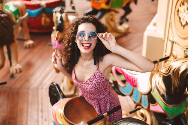 Lächelnde Dame mit dunklem lockigem Haar in Sonnenbrille und Kleid, die mit Lolly-Pop-Süßigkeiten in der Hand steht und glücklich in die Kamera schaut, während sie auf Karussell im Vergnügungspark fährt