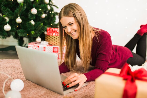 Lächelnde Dame mit Laptop nahe Geschenkboxen, Lichterketten und Weihnachtsbaum