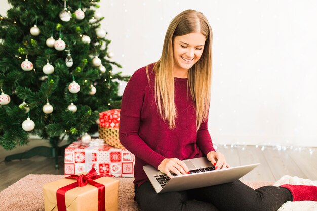 Lächelnde Dame mit Laptop nahe Geschenkboxen und Weihnachtsbaum