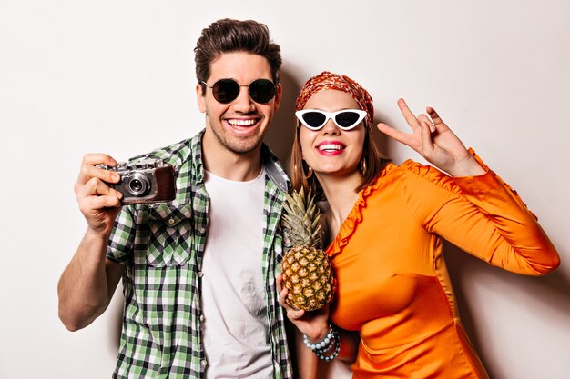 Lächelnde Dame mit rotem Lippenstift im orangefarbenen Kleid zeigt Friedenszeichen und hält Ananas. Kerl in der Sonnenbrille lächelt und macht Fotos auf Retro-Kamera.