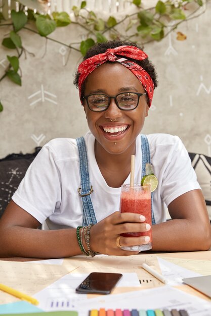 Lächelnde dunkelhäutige modische Frau mit erfreutem Ausdruck, genießt frischen Smoothie, arbeitet mit Dokumentation