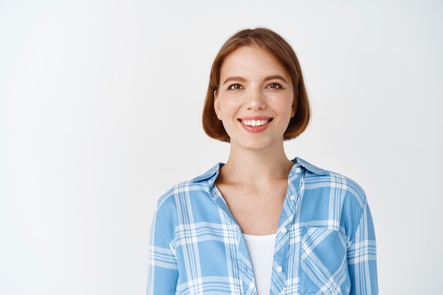 Lächelnde ehrliche Frau in Freizeitkleidung, die glücklich aussieht. Junges Mädchen mit kurzen Haaren und hellem Make-up, das auf weißer Wand steht