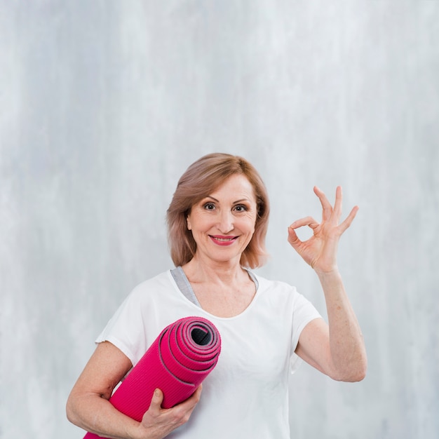 Lächelnde Eignungsfrau, die Yogamatte hält und okayzeichen mit den Fingern gegen graue Wand zeigt