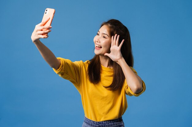 Lächelnde entzückende asiatische Frau, die Selfie-Foto auf Smartphone mit positivem Ausdruck in lässiger Kleidung macht und lokalisiert auf blauem Hintergrund steht. Glückliche entzückende frohe Frau freut sich über Erfolg.