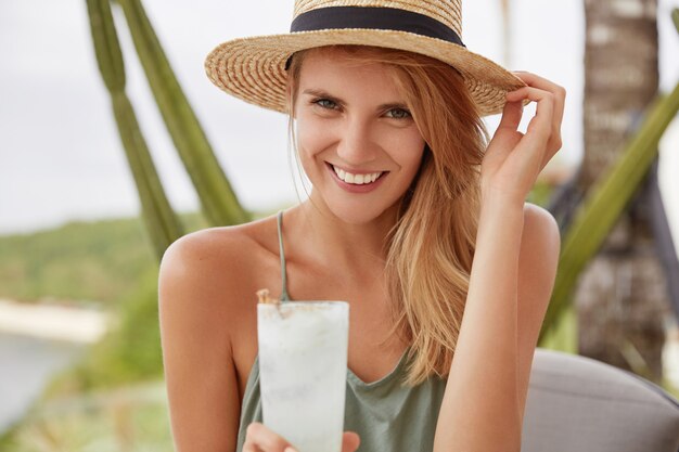 Lächelnde entzückende Frau mit glücklichem Ausdruck hat Sommerferien, verbringt Freizeit im Straßencafé mit frischem kaltem Getränk, sieht positiv aus. Attraktive Frau im Strohhut mit guter Laune.