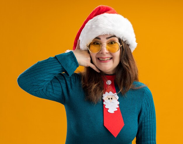 Lächelnde erwachsene kaukasische Frau in Sonnenbrille mit Weihnachtsmütze und Weihnachtskrawattengestikulieren rufen mich Zeichen lokalisiert auf orange Hintergrund mit Kopienraum