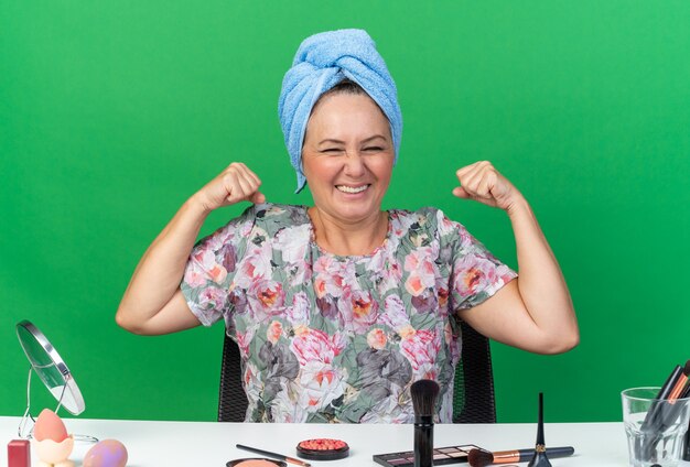 Lächelnde erwachsene kaukasische Frau mit eingewickeltem Haar in Handtuch, die am Tisch mit Make-up-Tools sitzt und ihren Bizeps anspannt, isoliert auf grüner Wand mit Kopierraum