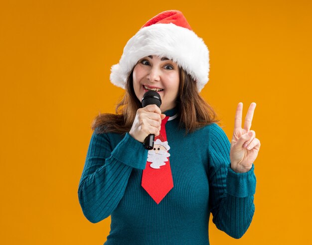 lächelnde erwachsene kaukasische Frau mit Santa Hut und Santa Krawatte, die Mikrofon und gestikulierendes Siegeszeichen lokalisiert auf orange Hintergrund mit Kopienraum hält