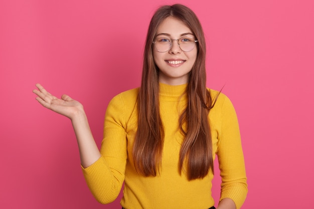 Lächelnde europäische Frau mit charmantem Gesichtsausdruck, der gelbes Freizeithemd und eine Brille trägt, Handfläche beiseite spreizend, isoliert über Rosenwand stehend.