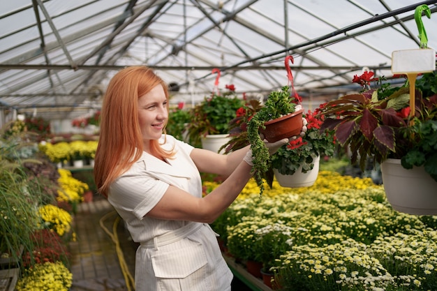 Lächelnde Floristin in ihrem Kinderzimmer, die Topfblumen inspiziert, während sie sich um die Gartenpflanzen im Gewächshaus kümmert