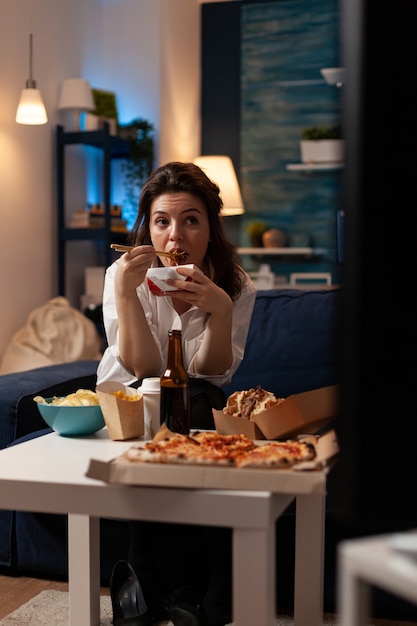 lächelnde Frau, die auf der Couch sitzt und leckeres chinesisches Essen isst, während Fastfood nach Hause geliefert wird?