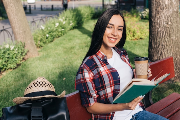 Lächelnde frau, die buch und wegwerfkaffeetasse beim sitzen auf bank am park hält