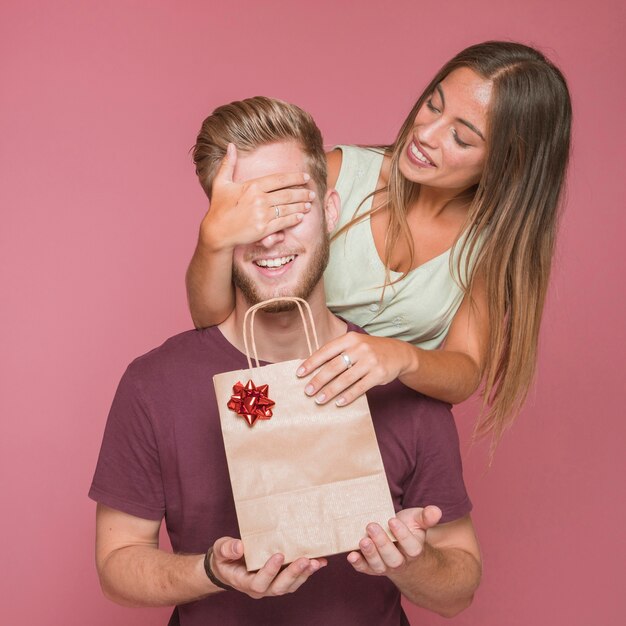 Lächelnde Frau, die das Auge ihres Freundes ihm Geschenkeinkaufstasche gebend bedeckt