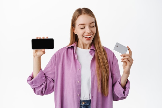 Lächelnde Frau, die glücklich auf ihre Kreditkarte schaut, einen horizontalen Smartphone-Bildschirm zeigt, eine Anwendung oder einen Internet-Shop empfiehlt und über der weißen Wand steht