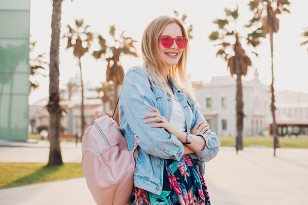 lächelnde Frau, die in der Stadtstraße im stilvollen bedruckten Rock und in der übergroßen Jeansjacke mit rosa Sonnenbrille geht