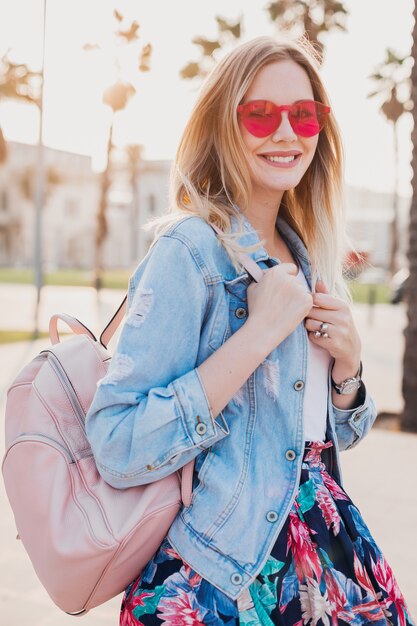 lächelnde Frau, die in der Stadtstraße im stilvollen bedruckten Rock und in der übergroßen Jeansjacke mit rosa Sonnenbrille geht