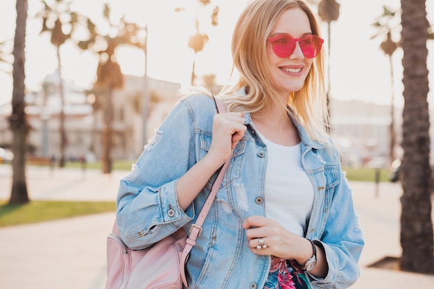 lächelnde Frau, die in der Stadtstraße in stilvoller Jeans-Oversize-Jacke mit rosa Sonnenbrille und Lederrucksack geht
