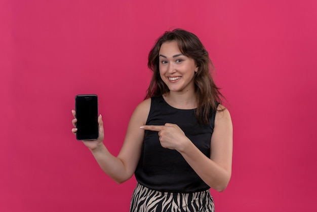 Lächelnde Frau, die schwarzes Unterhemd hält, das ein Telefon hält und das Telefon auf rosa Wand zeigt