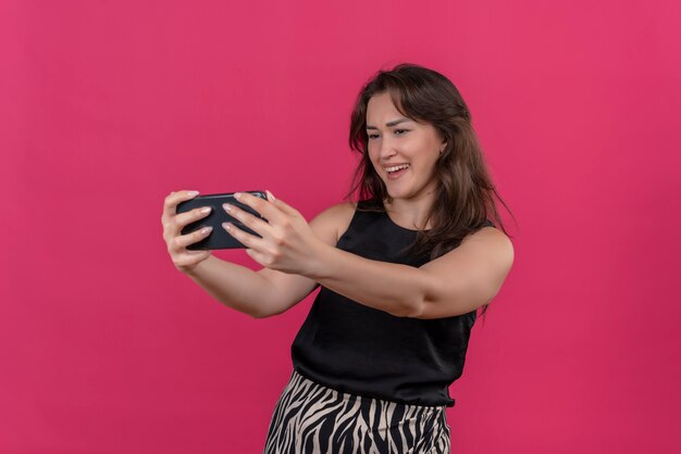 Lächelnde Frau, die schwarzes Unterhemd trägt, nehmen ein Selfie auf rosa Wand