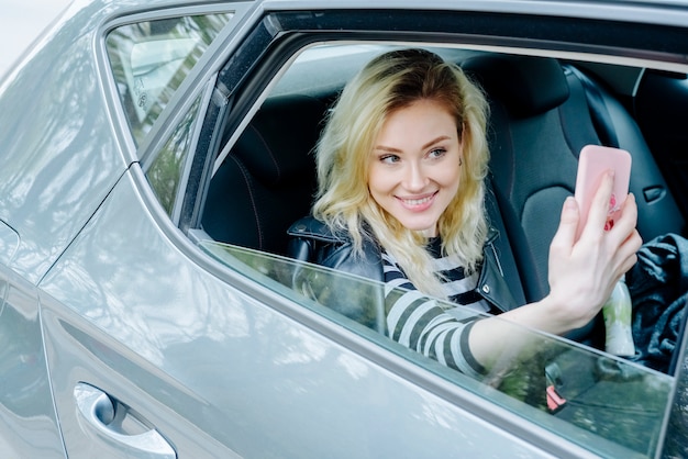 Lächelnde Frau, die selfie mit Smartphone nimmt, während sie in einem Auto in der schwarzen Lederjacke und im gestreiften Oberteil sitzt