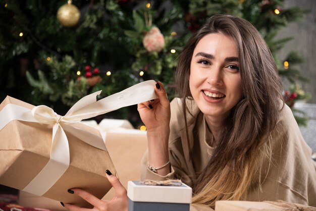 Lächelnde Frau, die sich auf einem flauschigen Teppich hinlegt und ein Weihnachtsgeschenk einwickelt.