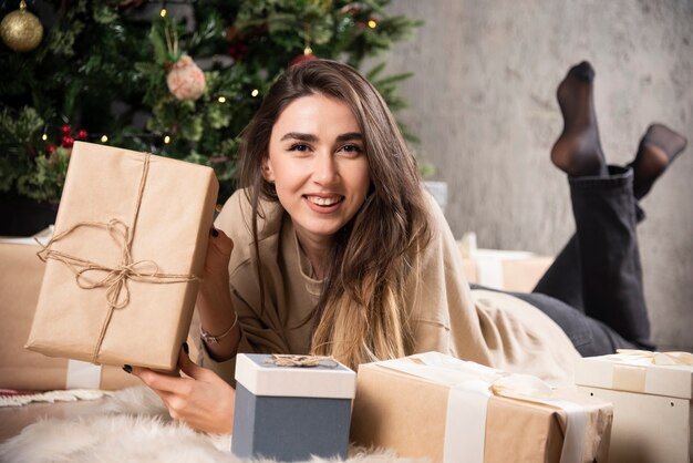 Lächelnde Frau, die sich auf flauschigem Teppich hinlegt und ein Weihnachtsgeschenk zeigt.