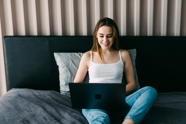 Lächelnde Frau holt ihre sozialen Medien ein, während sie sich an einem faulen Tag mit einem Laptop im Bett entspannt
