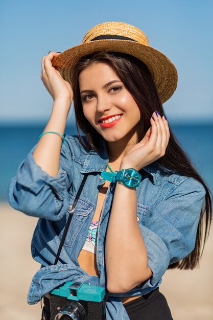 Lächelnde Frau im Strohhut und im stilvollen Sommeroutfit, das mit Retro-Kamera am Strand aufwirft.