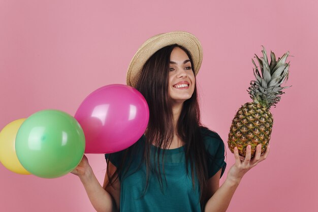 Lächelnde Frau in einem Hut wirft mit bunten Ballonen und einer Ananas auf