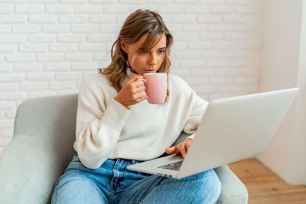 Lächelnde Frau mit blonden, gewellten Haaren, die zu Hause auf dem Sofa sitzt, an einem Laptop arbeitet und eine Tasse Kaffee hält