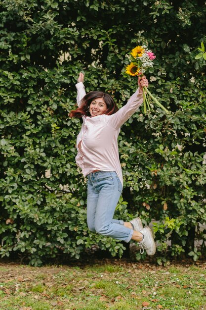 Lächelnde Frau mit Blumenstrauß springend nahe grünem Busch