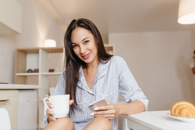 Lächelnde Frau mit den schwarzen Haaren, die in der Küche mit Smartphone während des Frühstücks sitzen
