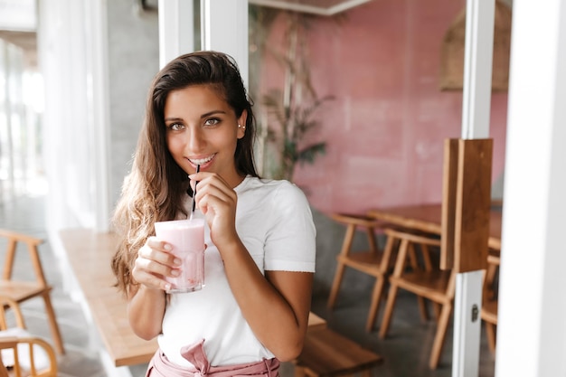 Lächelnde Frau mit grünen Augen in weißem T-Shirt, die Milchshake im Café trinkt