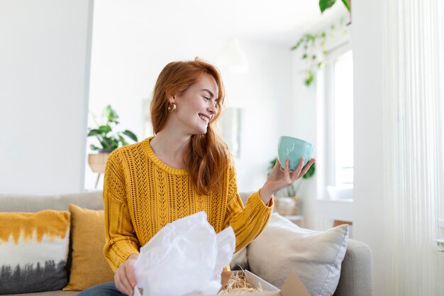 Lächelnde Frau sitzt zu Hause auf der Couch und kauft online Waren im Internet. Glückliche junge Kundin packt das Postversandpaket aus, das mit der Bestellung oder Lieferung zufrieden ist