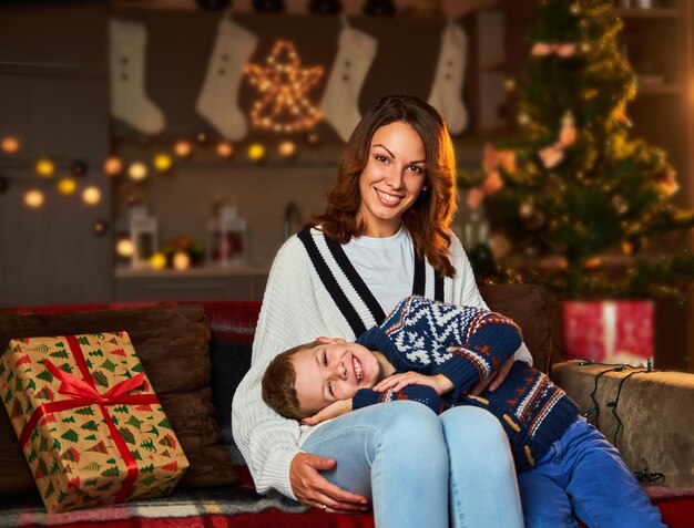 Lächelnde Frau und kleiner Junge, die in einem Wohnzimmer mit weihnachtlich beleuchtetem Hintergrund posieren.