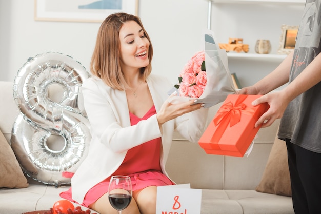 Lächelnde Frauen, die am glücklichen Frauentag auf dem Sofa sitzen, geben von jemandem im Wohnzimmer ein Geschenk mit Blumenstrauß