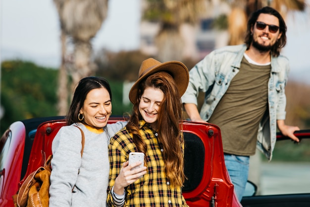 Lächelnde Frauen, die selfie auf Smartphone nahe dem Autostiefel nehmen und Mann, der sich heraus vom Auto lehnt