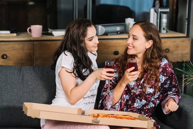 Lächelnde Freundinnen, die auf Sofa mit Getränken und Pizza sitzen