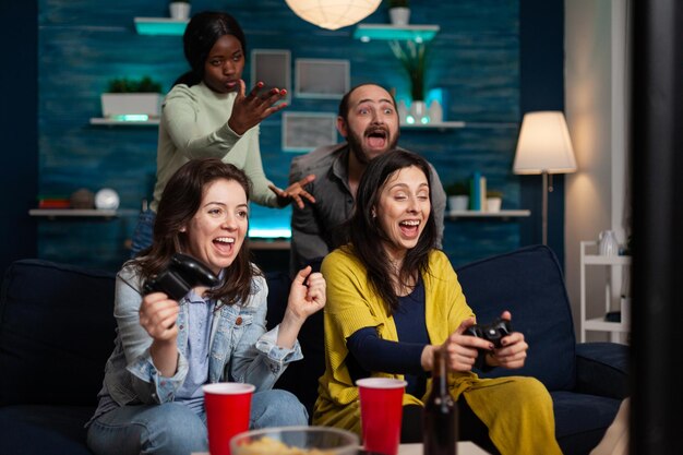 Lächelnde Freundinnen sitzen auf der Couch und gewinnen Videospiele mit Spielkonsolen, die während des Online-Wettbewerbs Spiele spielen. Multiethnische Gruppe von Menschen, die gerne Zeit miteinander verbringen. Freundschaftskonzept