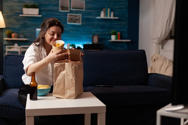 Lächelnde fröhliche frau beim auspacken von fast-food-mahlzeiten auf dem sofa während junkfood nach hause geliefert