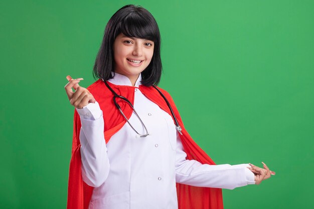 Lächelnde, gerade aussehende junge Superheldenmädchen tragend Stethoskop mit medizinischem Gewand und Umhang lokalisiert auf Grün