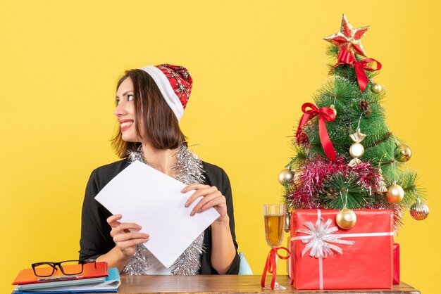 Lächelnde Geschäftsdame im Anzug mit Weihnachtsmannhut und Neujahrsdekorationen, die allein arbeiten und an einem Tisch mit einem Weihnachtsbaum darauf im Büro sitzen